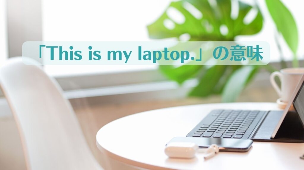 丸い形のデスクの上にスマートホンとラップトップパソコン、マグカップなどがあります。デスクの向こうには観葉植物が見えます。「This is my laptop.」という英文の意味を説明するための画像です。