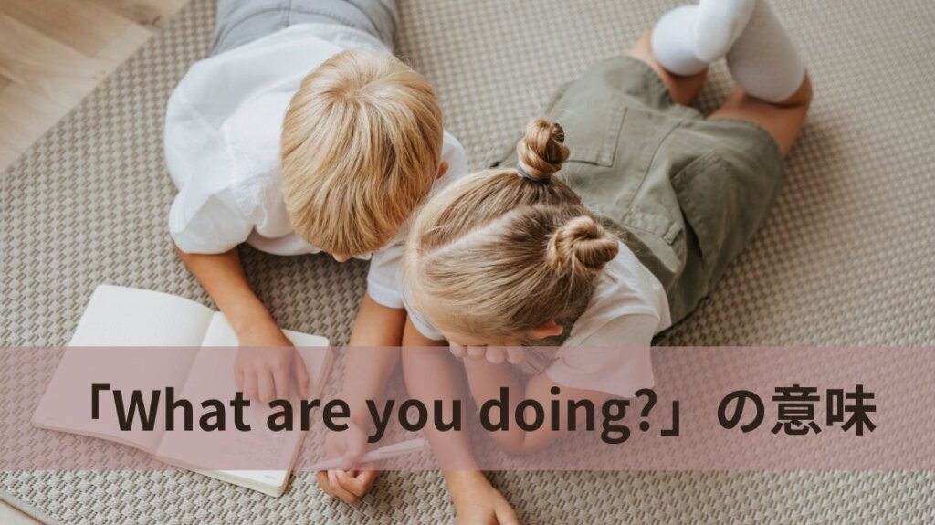 金髪に白いシャツの子どもが二人、床に腹ばいに寝そべってノートに何かを書きこんでいいます。「What are you doing?」という文の説明のための画像です。
