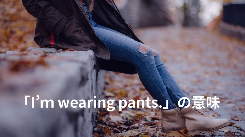デニムパンツとジャケットに身を包んだ女性の胸から下の写真です。季節は秋から冬らしく石垣に腰を掛けている女性の足元には枯れ葉が積み重なっています。「I’ｍ wearing pants.」という英語の説明のためにデニムパンツをはいている人の写真を使いました。