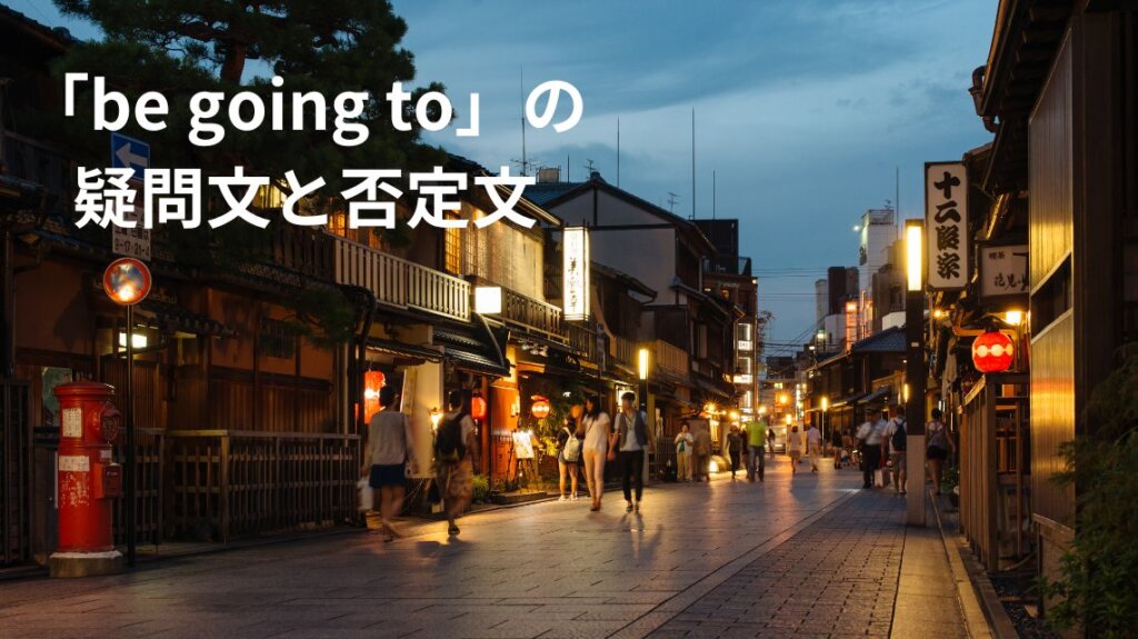 夜の京都の街並みです。左には昔ながらの赤いポストがあります。建物は木造二階建てで、看板やちょうちんを飾っているところもあります。真ん中の道を観光客が往来しています。